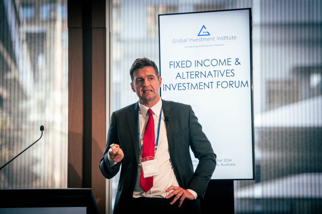 A professional speaker presenting at the Fixed Income & Alternatives Investment Forum, with a screen displaying the event title in the background.