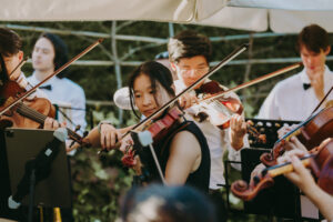 Group of musicians performing outdoors with violins, dressed in formal attire under a tent.