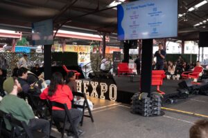 Outdoor HEXPO stage under a covered market pavilion with a speaker presenting to a seated audience, large screens and illuminated HEXPO sign.