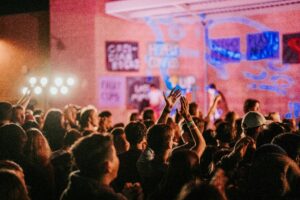 Packed crowd at an urban warehouse concert with hands raised, stage lighting and projection on a brick wall.