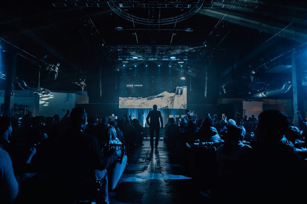 Crowd facing a large LED stage in an industrial indoor venue with atmospheric blue lighting and overhead truss rigging.