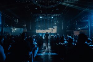 Crowd facing a large LED stage in an industrial indoor venue with atmospheric blue lighting and overhead truss rigging.
