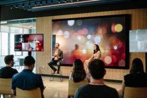 Hybrid panel discussion in a modern corporate event space with a large LED video wall behind speakers and side screens showing remote participants.