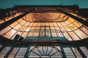 Glass-fronted Artvisory balcony and façade lit from warm interior chandeliers at dusk.